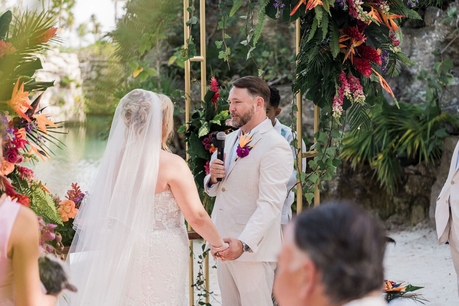 Couple exchanging vows at tropical outdoor wedding ceremony with colorful floral arch and lush greenery backdrop.