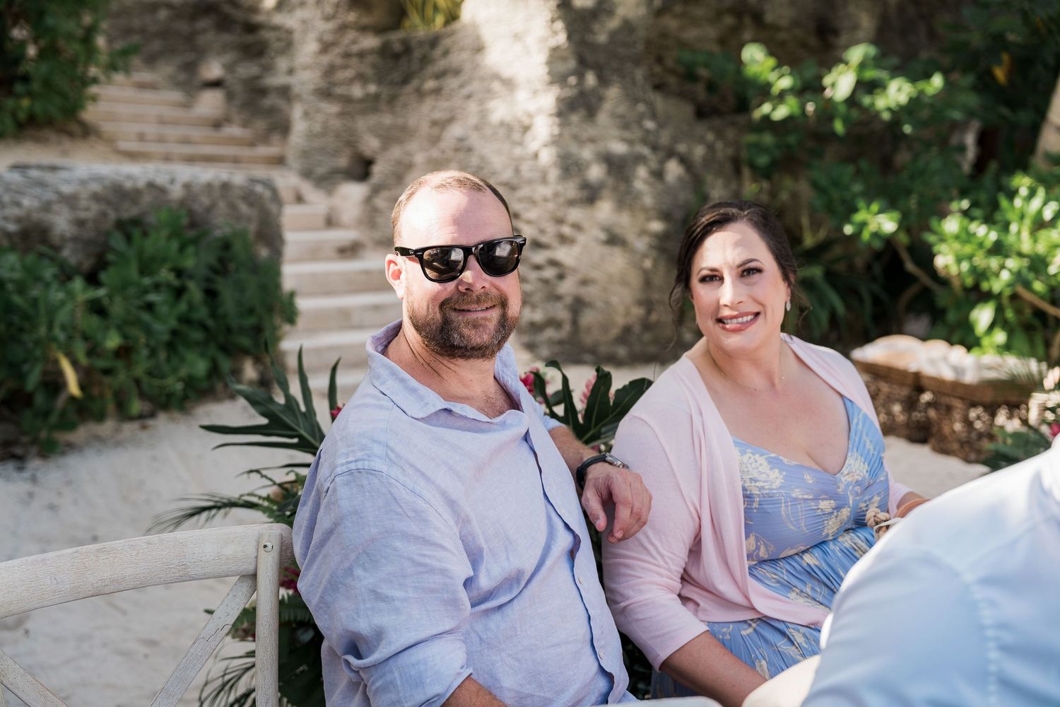 A smiling couple sits outdoors near stone steps and tropical plants, man wearing sunglasses in a blue shirt.