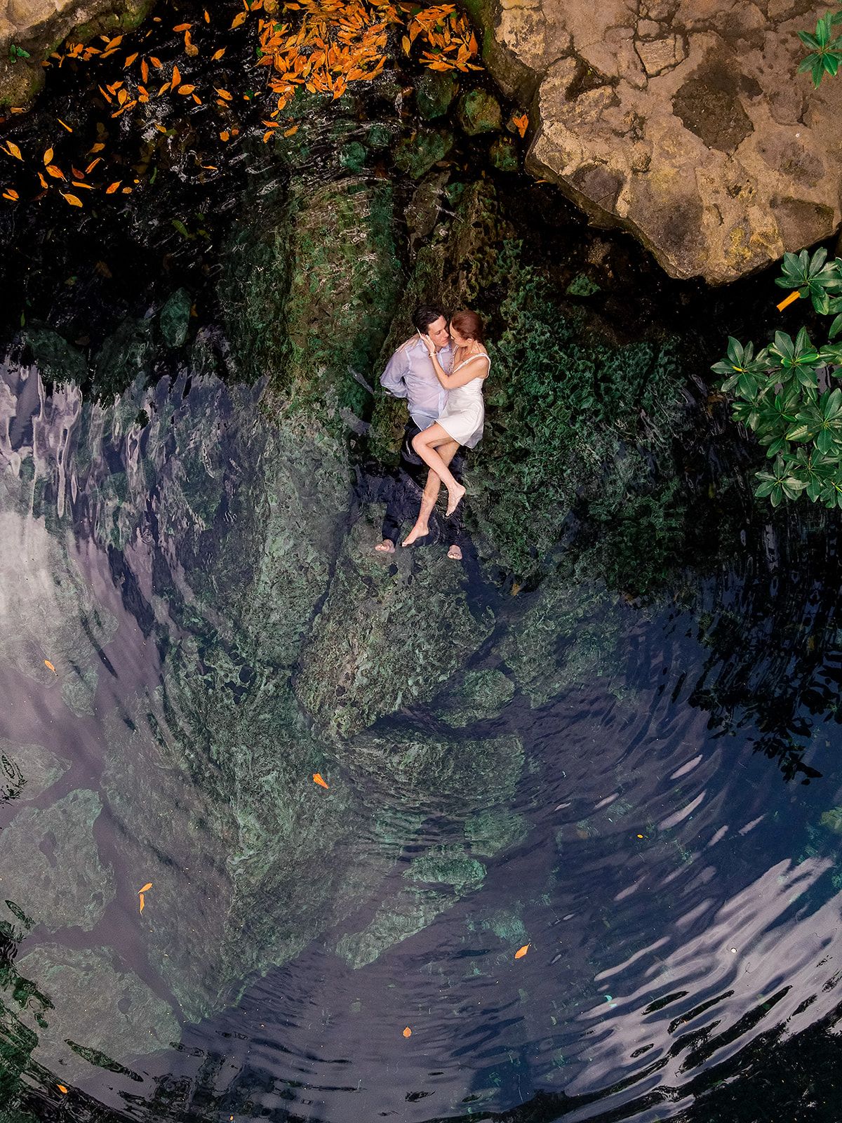 Aerial view of a romantic couple embracing on mossy rocks beside a tranquil cenote surrounded by lush greenery. Cenote photoshoot