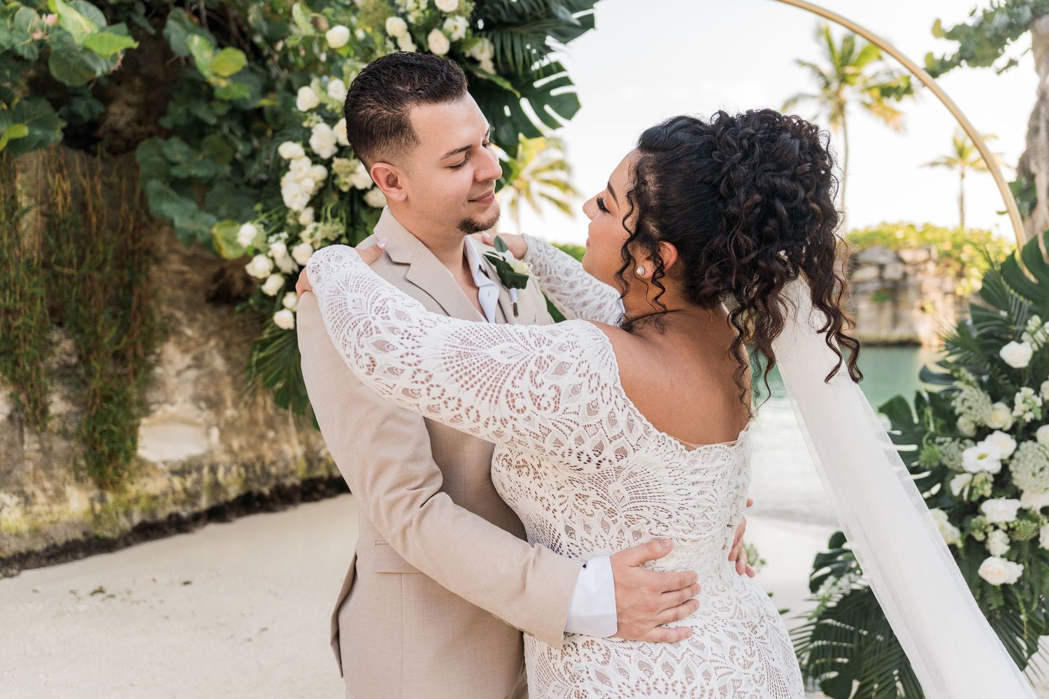Xcaret Arte Wedding - A couple shares a romantic embrace at their outdoor beach wedding, the groom in a tan suit and bride in a lace gown.