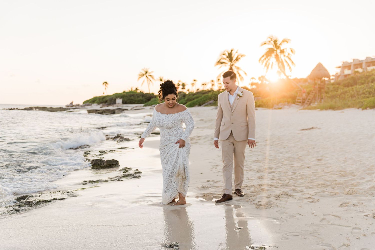 Xcaret Arte Wedding - Bride and groom walk hand-in-hand on a tropical beach at sunset with palm trees in the background.