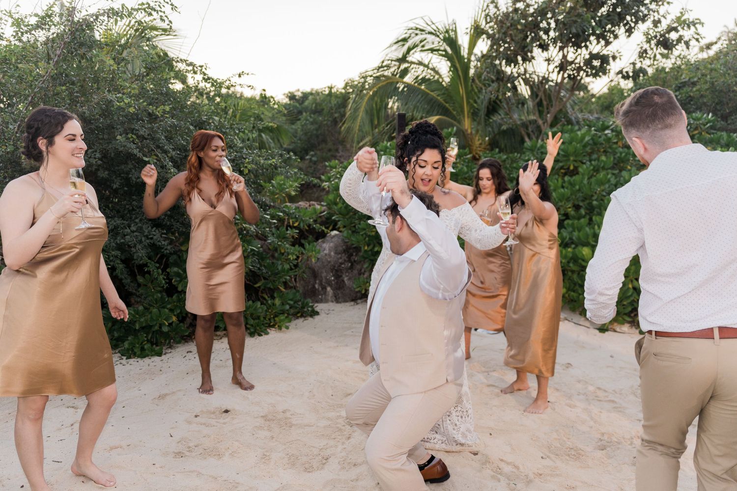 Xcaret Arte Wedding - Bride dances joyfully with wedding party on sandy beach surrounded by lush tropical greenery at sunset celebration.