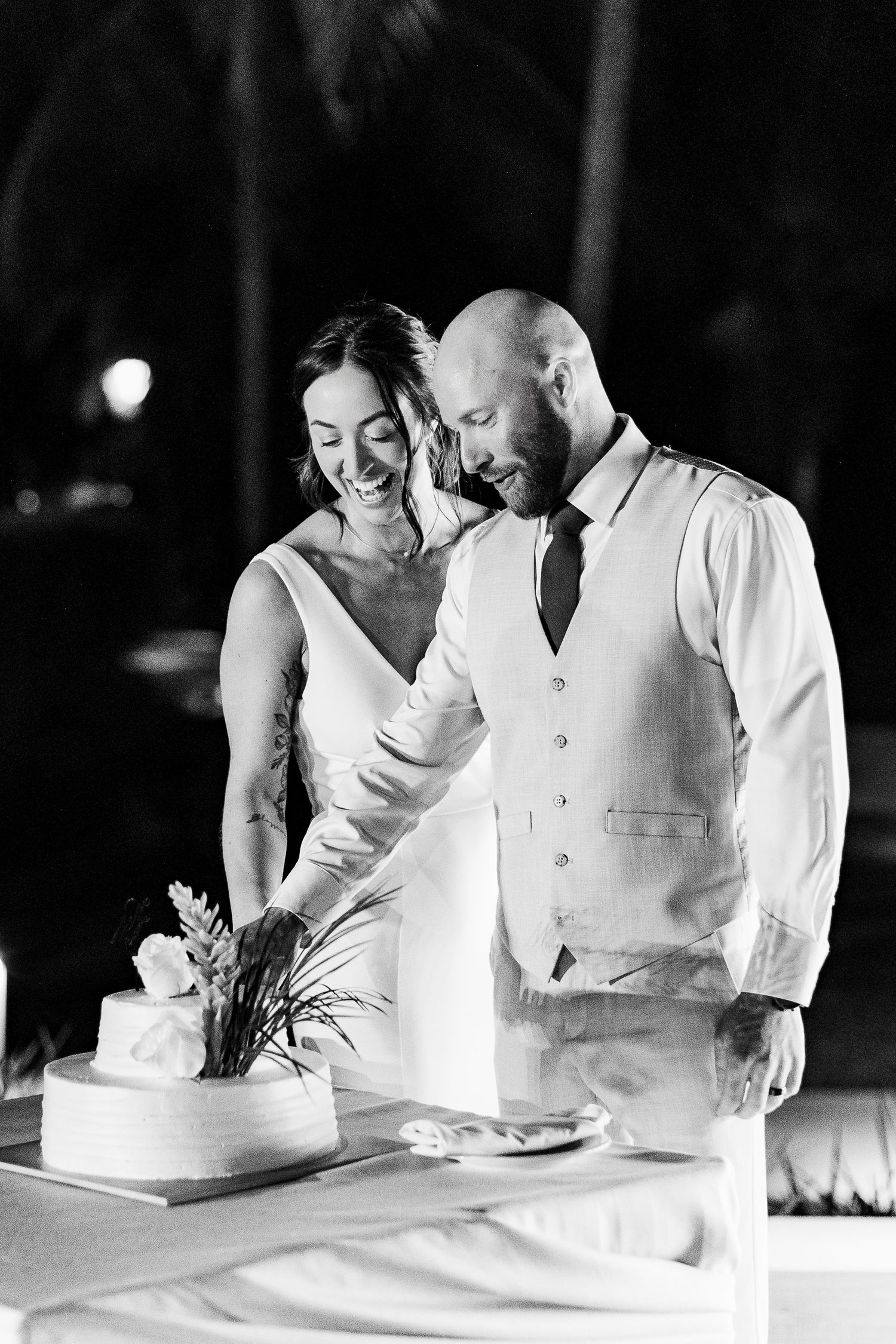 Black and white photo of a smiling bride and groom cutting their wedding cake at an outdoor evening reception.