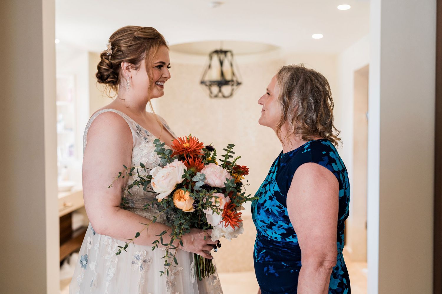Bride in floral gown holding colorful bouquet smiles with older woman in blue dress during wedding preparation.