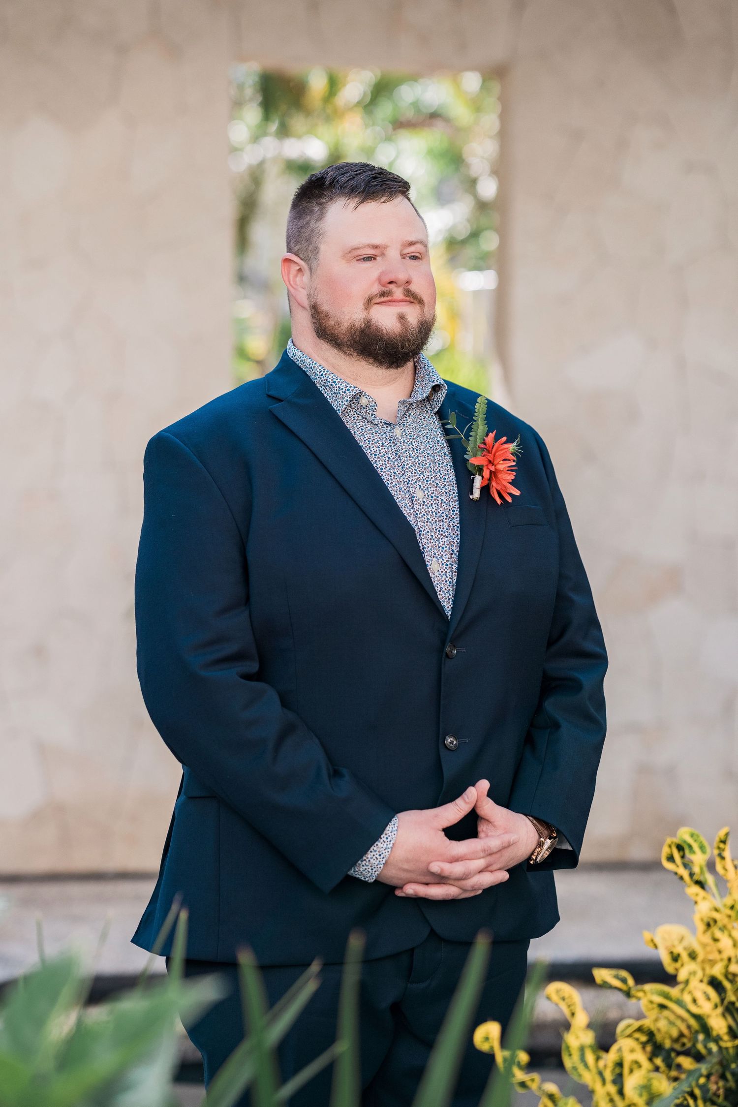 Secrets Akumal Wedding - Groom in navy suit with red boutonniere stands confidently outdoors at a wedding ceremony.