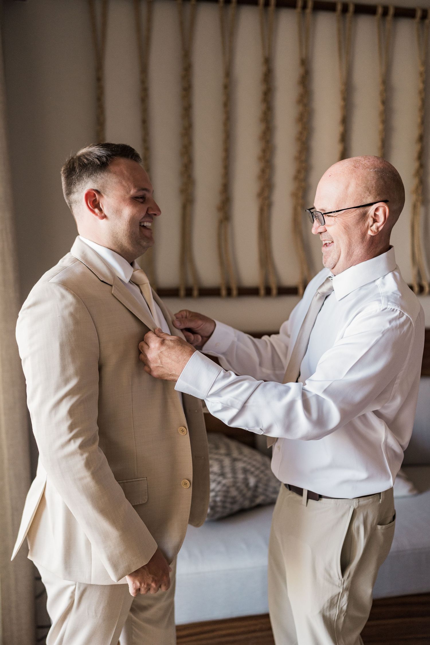 Groom in beige suit smiling as older man helps adjust his lapel during wedding preparations.