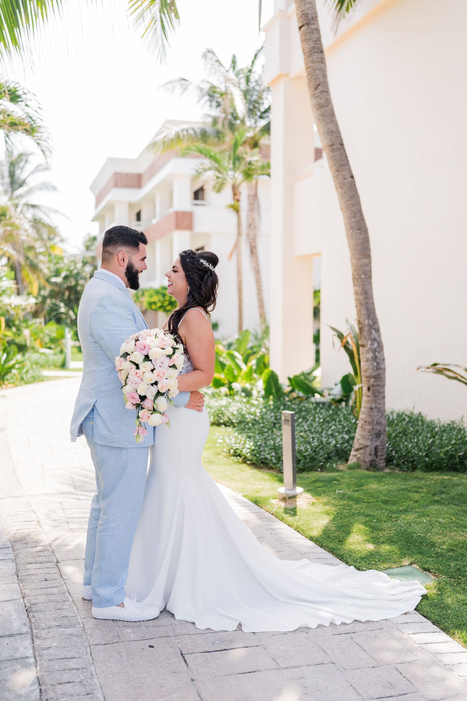 Bride and groom share romantic moment on tropical resort pathway holding pink and white bouquet.