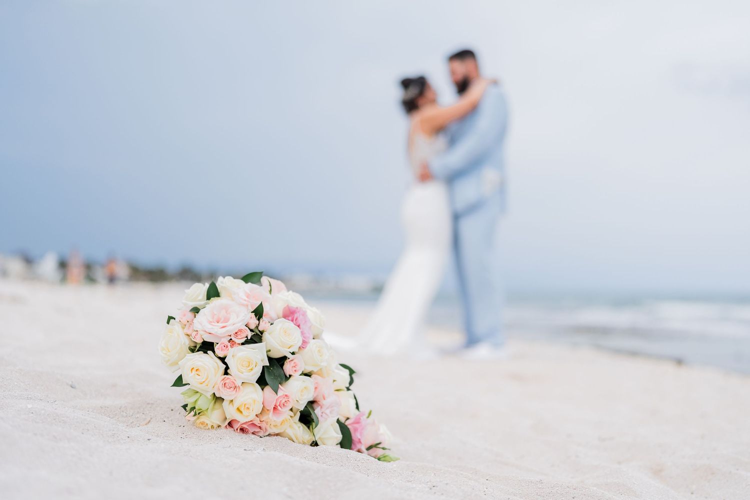 Pink and white bridal bouquet rests on white sand as couple embraces on the beach in the background.