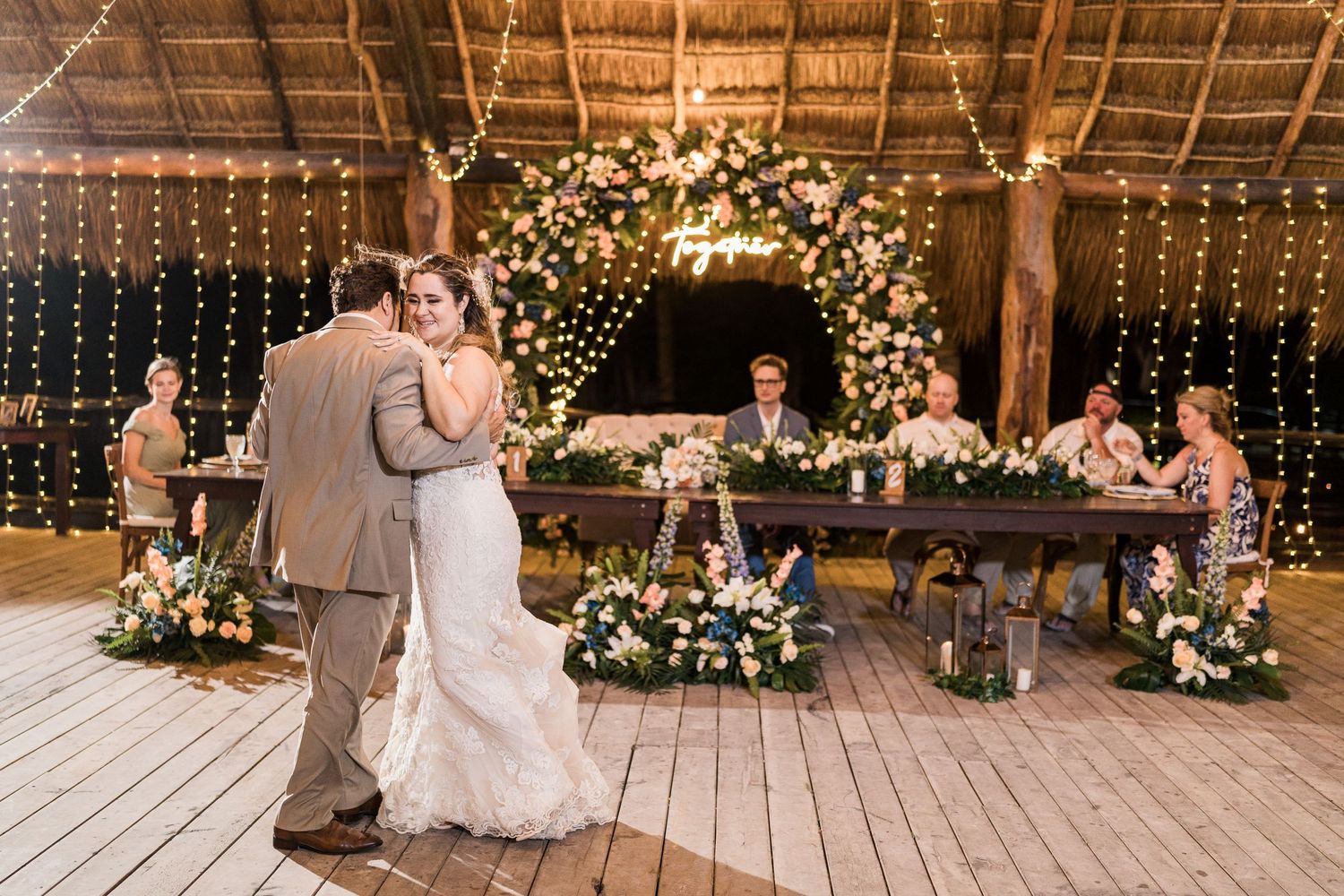 Couple dancing at wedding reception with floral arch and head table adorned with lush blooms behind them.