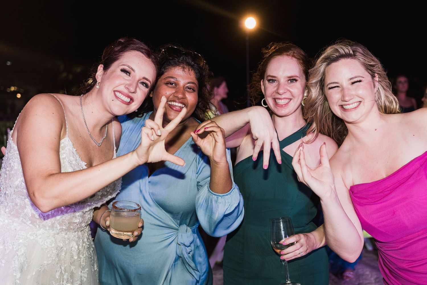 Four women in elegant dresses smile and pose together at a wedding reception celebration at night.