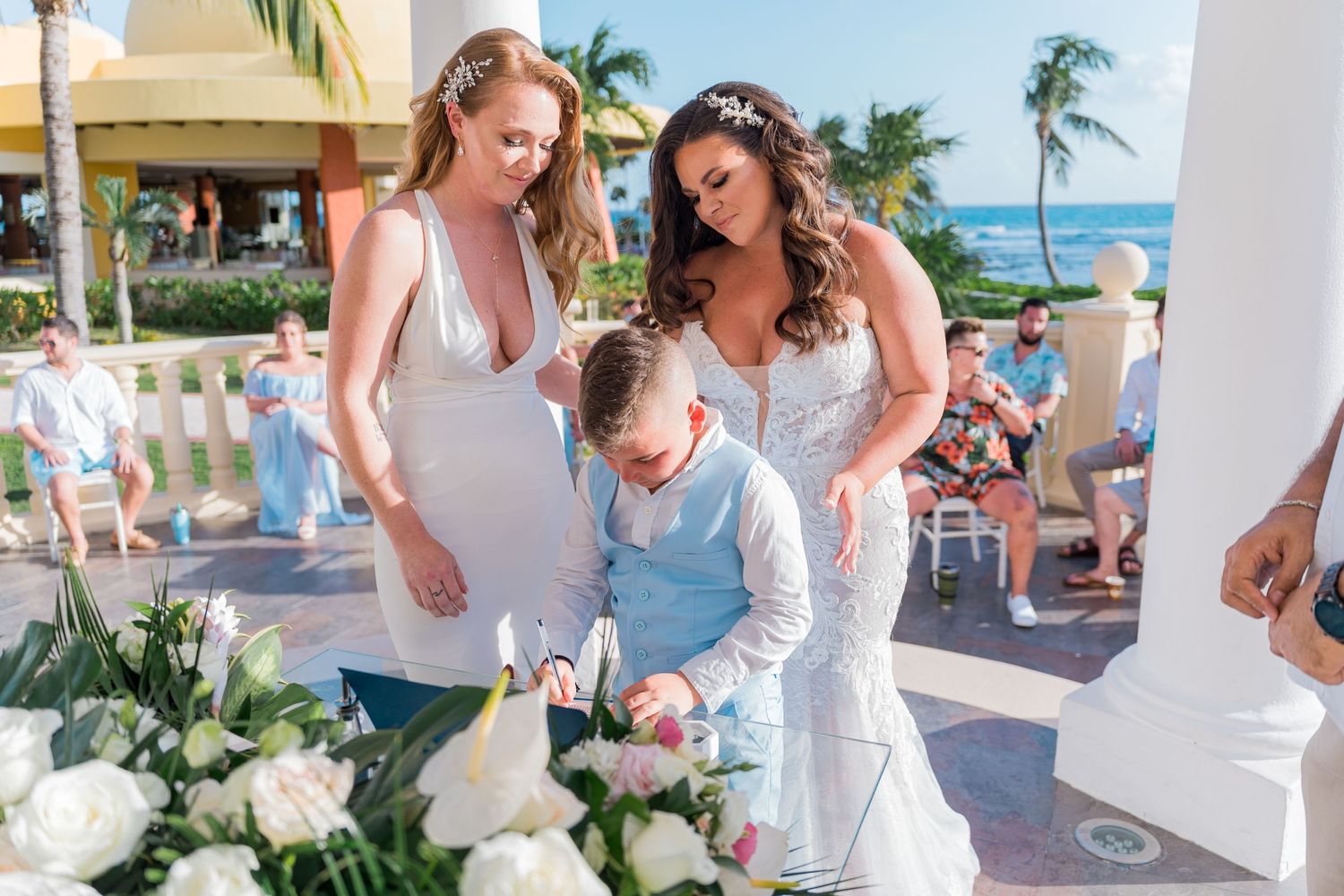 Two brides and a young boy signing the wedding register at a tropical outdoor ceremony.