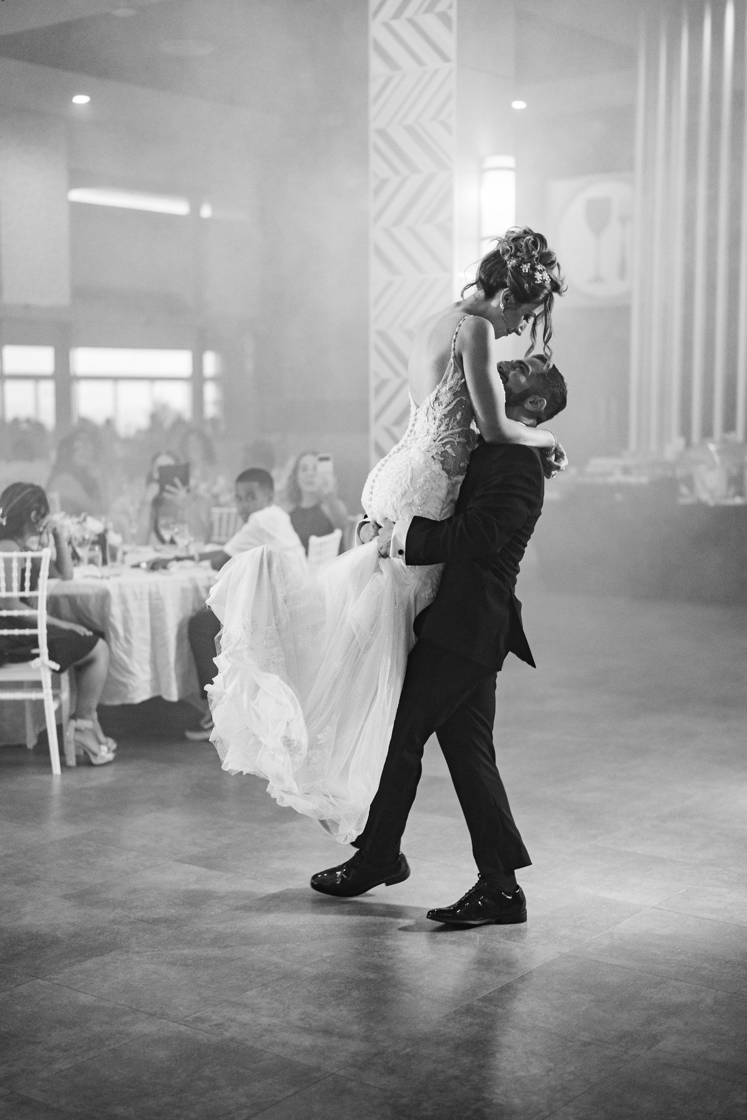 Black and white photo of groom lifting bride during emotional first dance at wedding reception.