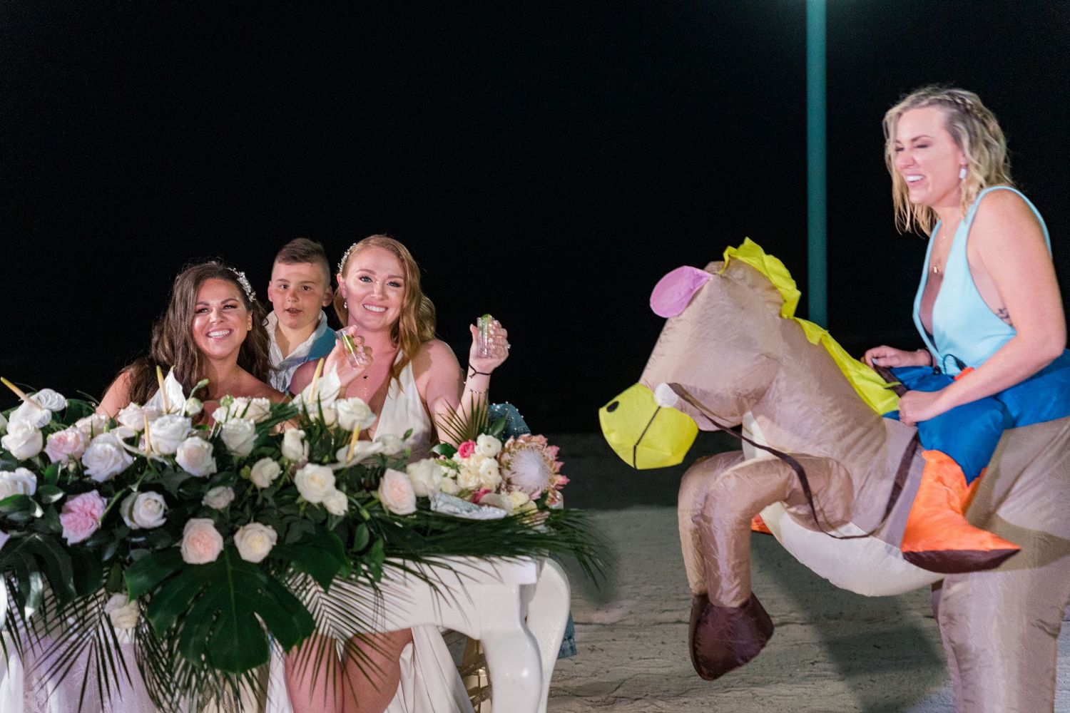 Bride and groom laugh as a wedding guest rides an inflatable horse costume at a beach reception.