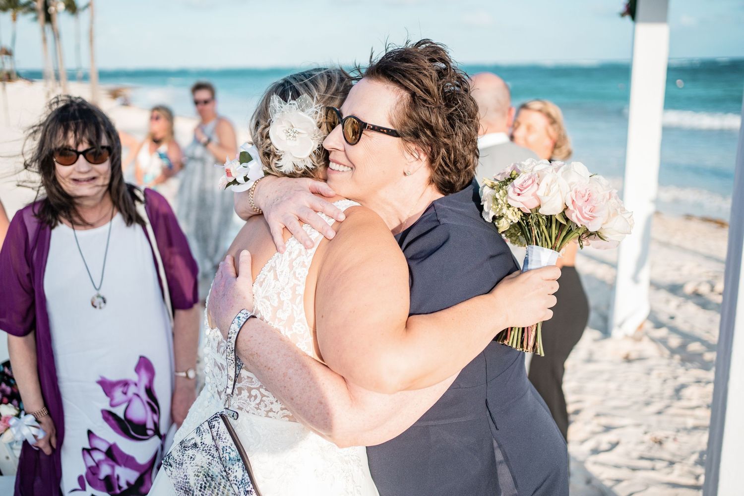 Bride in lace gown receives heartfelt hug from guest holding pink bouquet at beach wedding.