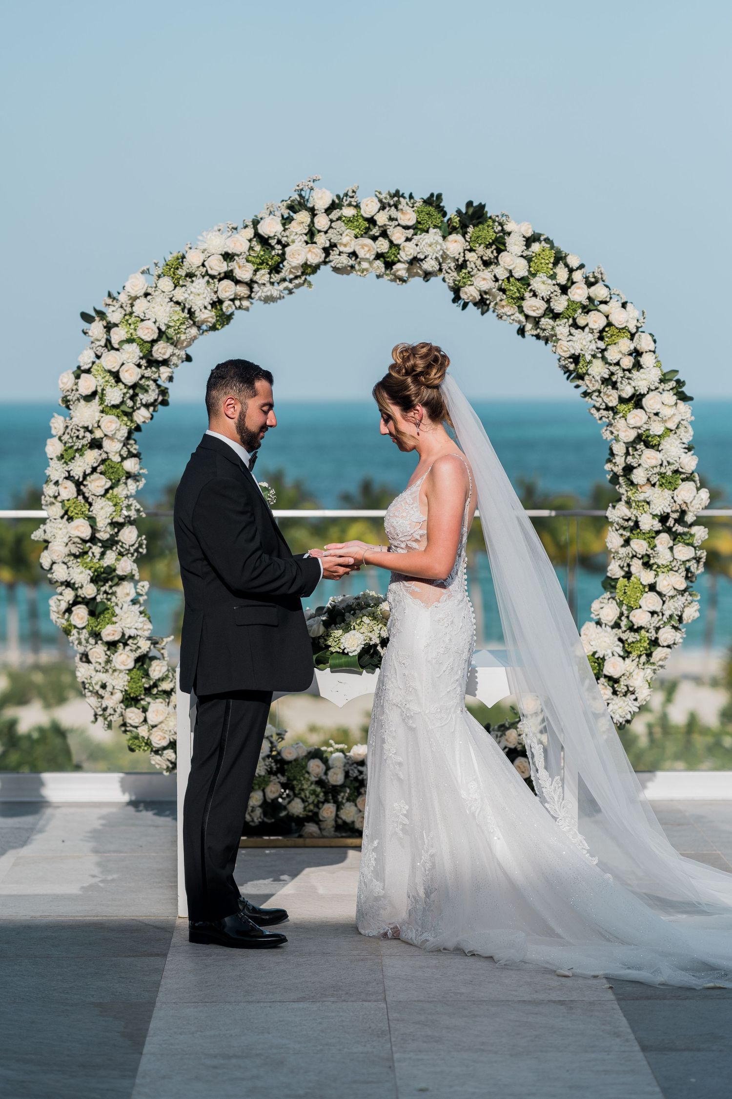 Couple exchanges rings under white floral circular arch at beachfront wedding ceremony.