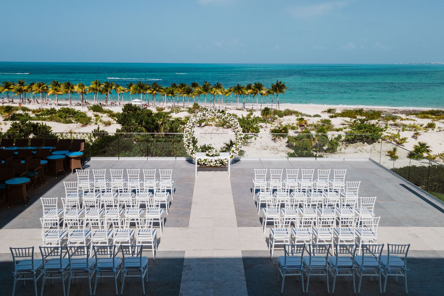 Aerial view of beachfront wedding ceremony setup with white chairs and floral arch.