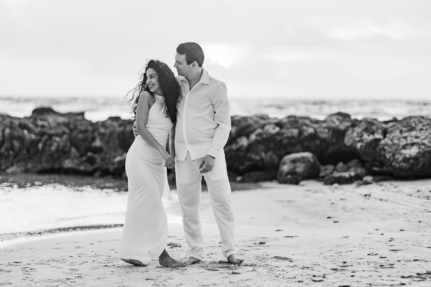 Black and white photo of a couple walking on a rocky beach, the woman in a white dress and man in white attire.