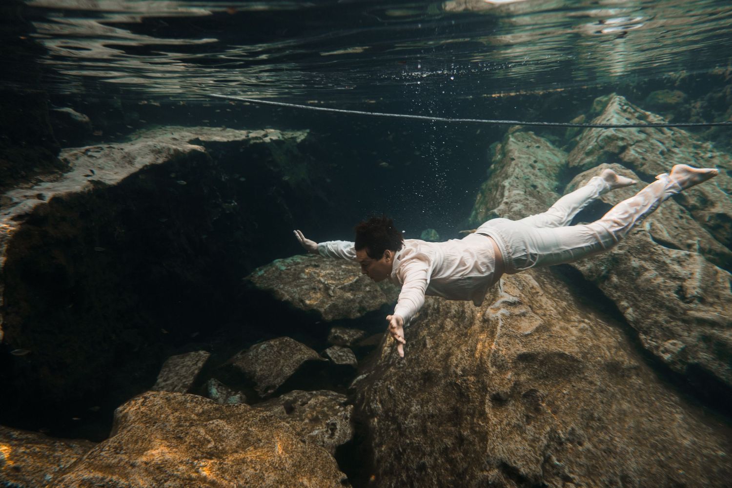 Person swimming underwater near rocky formations in clear water, wearing white clothing in a serene aquatic environment.