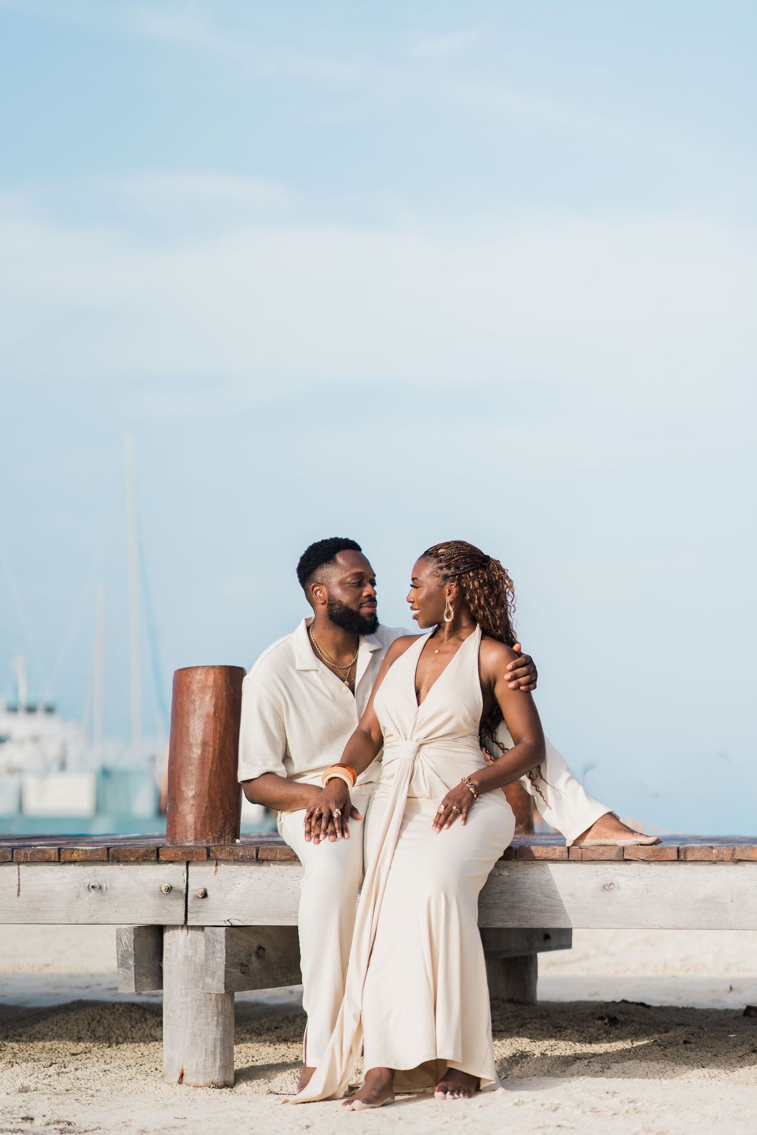 A couple in matching cream outfits sit together on a beach bench, gazing at each other near a marina.