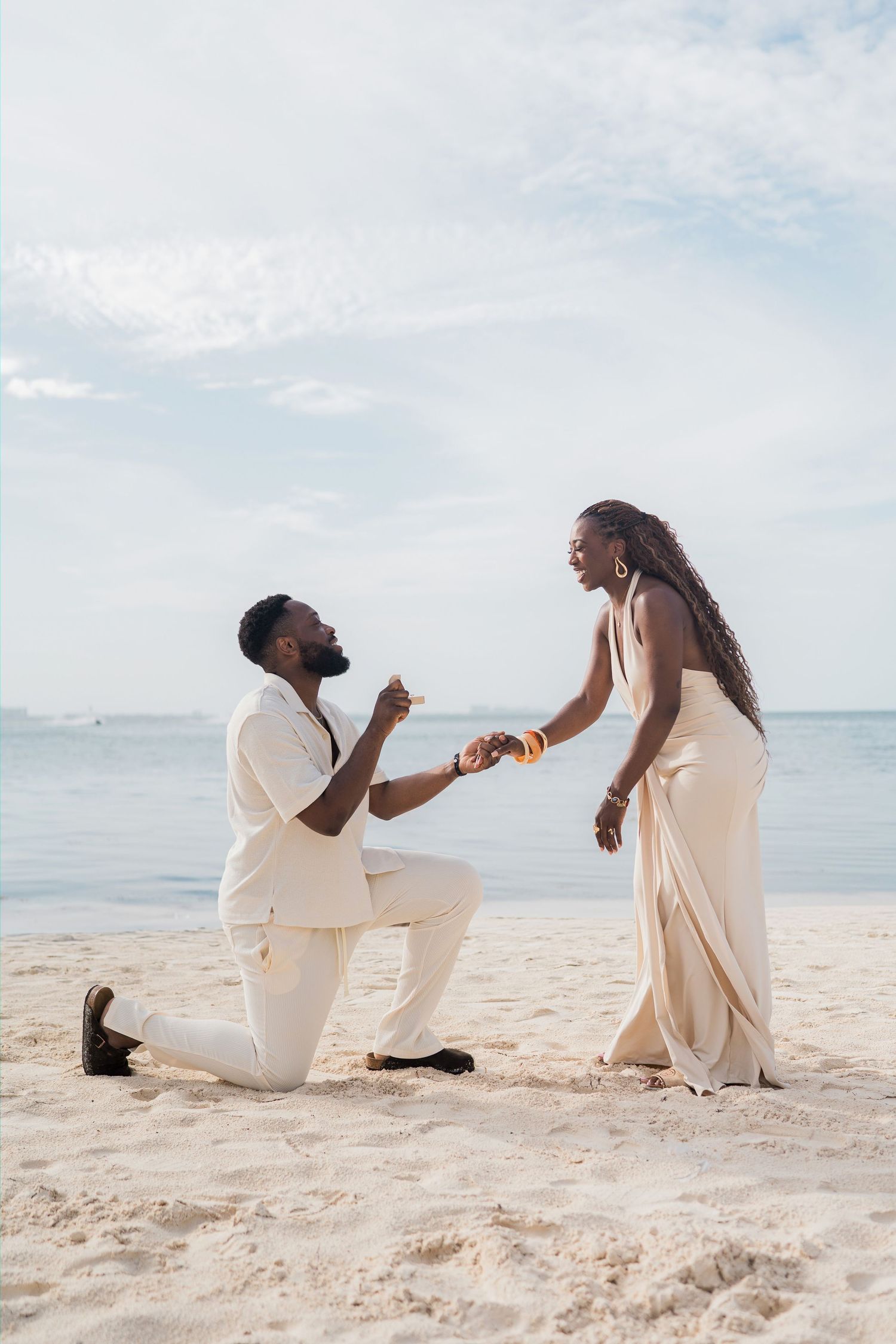 Man proposes to woman on one knee on a sandy beach, both dressed in elegant cream outfits with ocean backdrop.