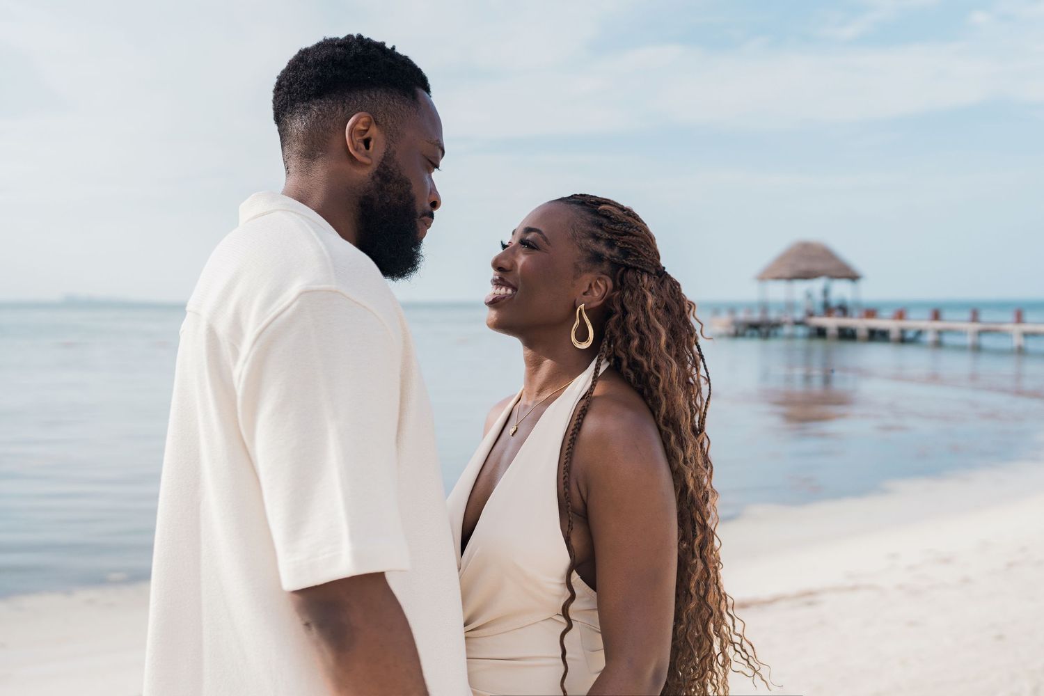 A couple in elegant cream attire sharing a romantic moment on a sunny beach with a pier behind them.