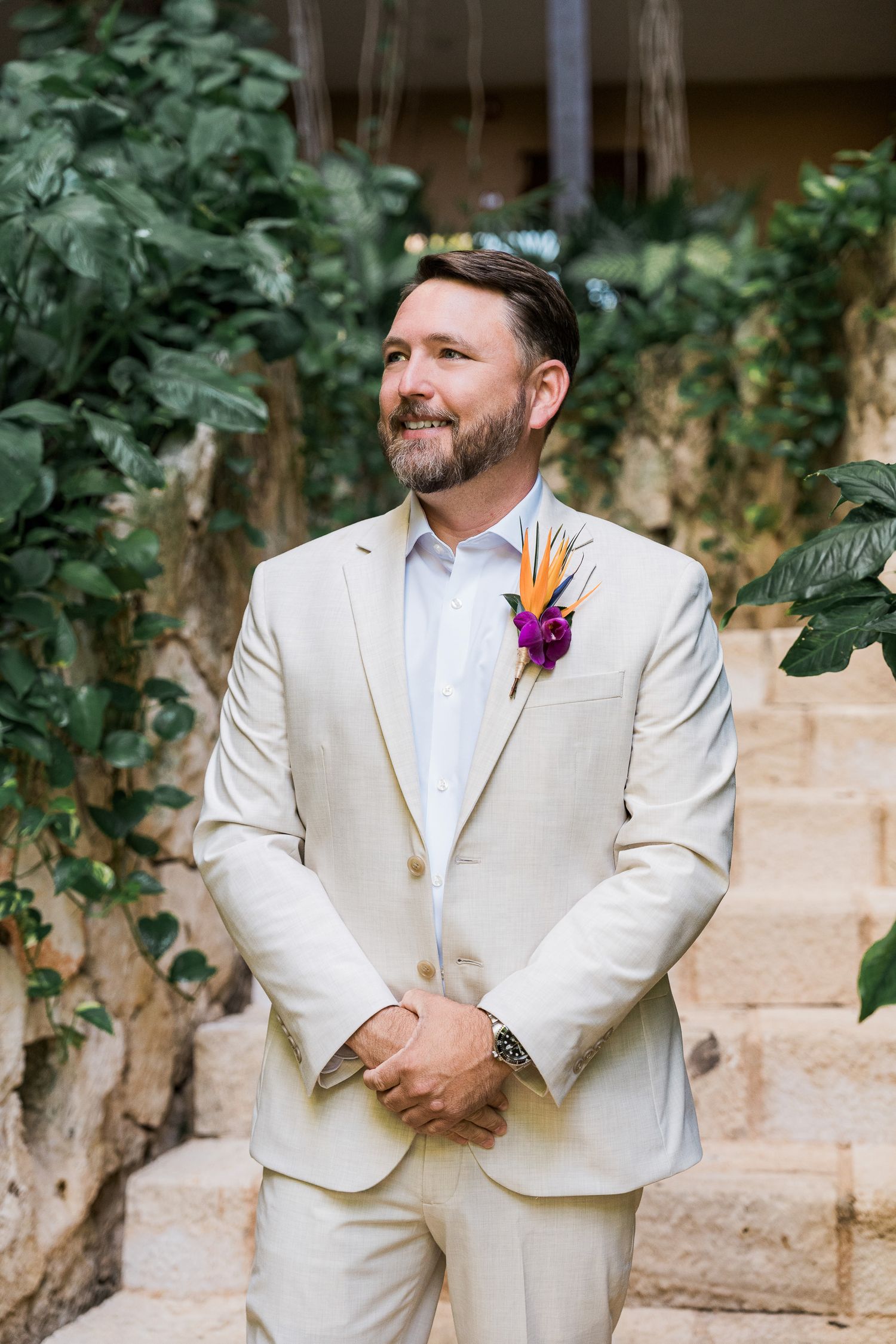 Groom in cream suit with tropical boutonniere smiling outdoors at a destination wedding.
