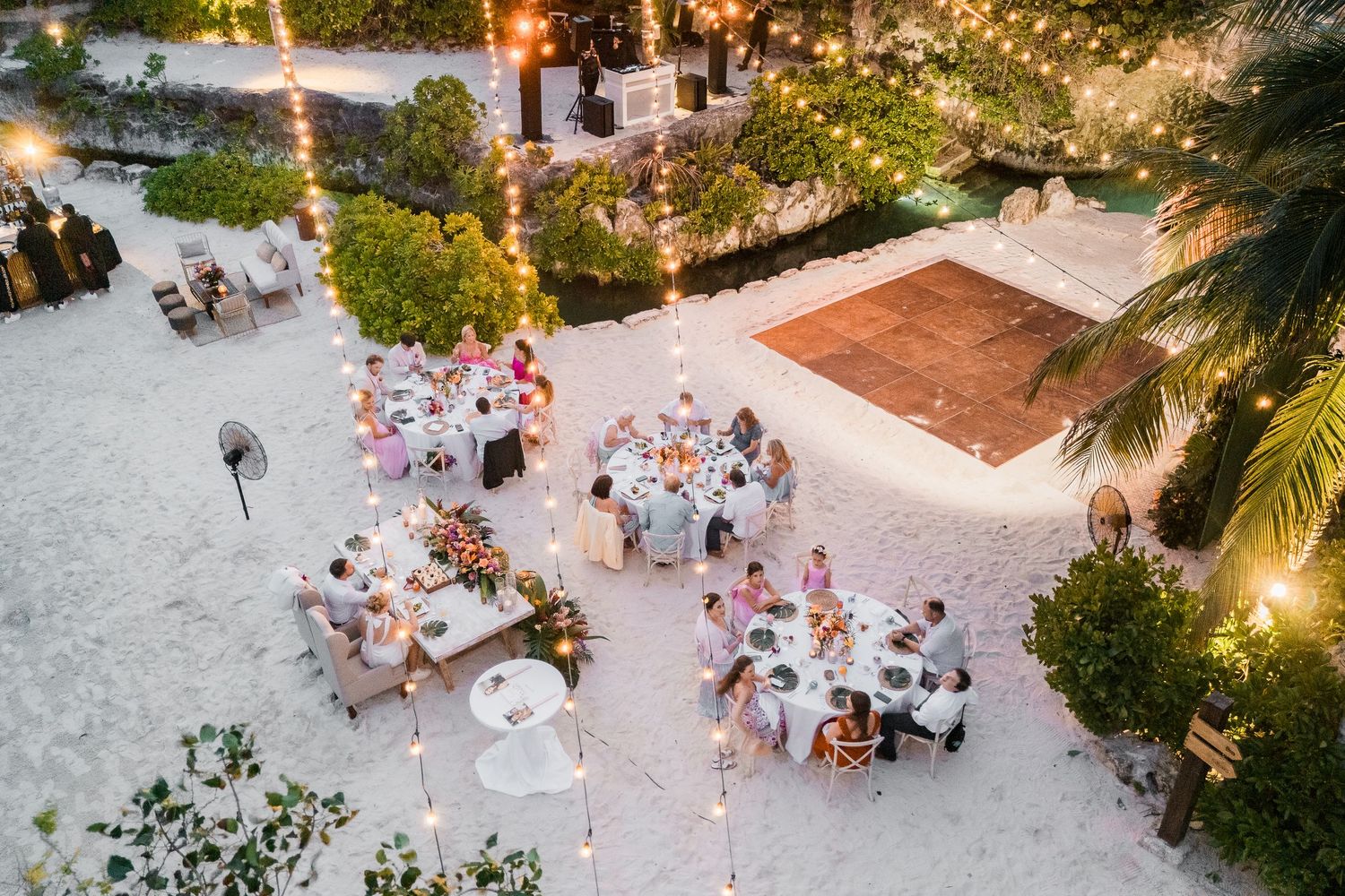 Hotel Xcaret México Aerial view of an elegant outdoor wedding reception with string lights, round tables, and a dance floor on white sand.