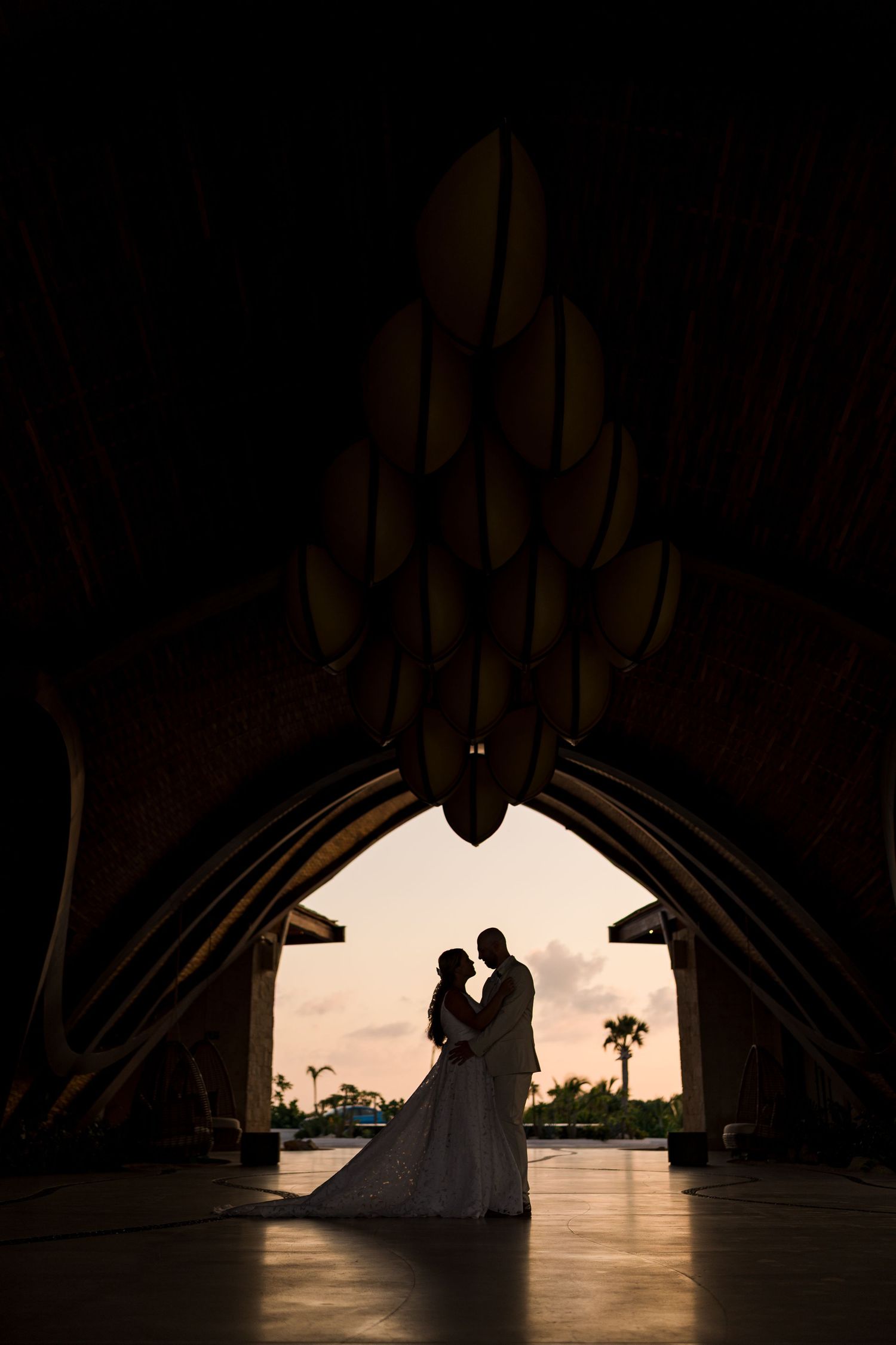 Silhouette of bride and groom embracing under dramatic arched venue entrance at sunset.