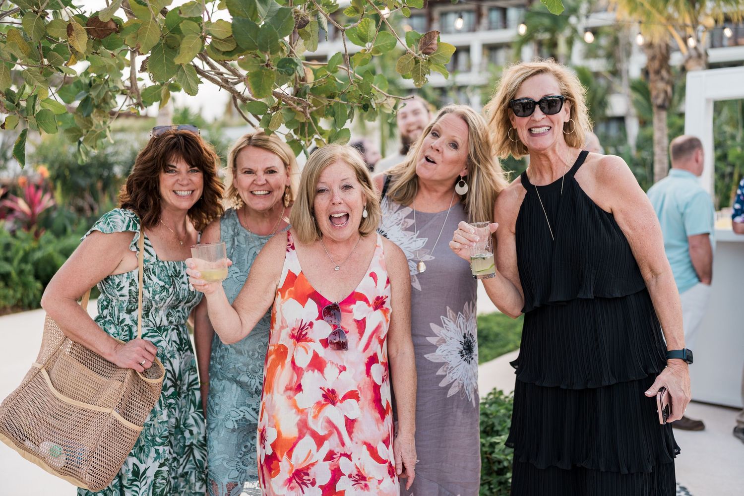 Five women smiling and posing together outdoors at a tropical resort during a social gathering.