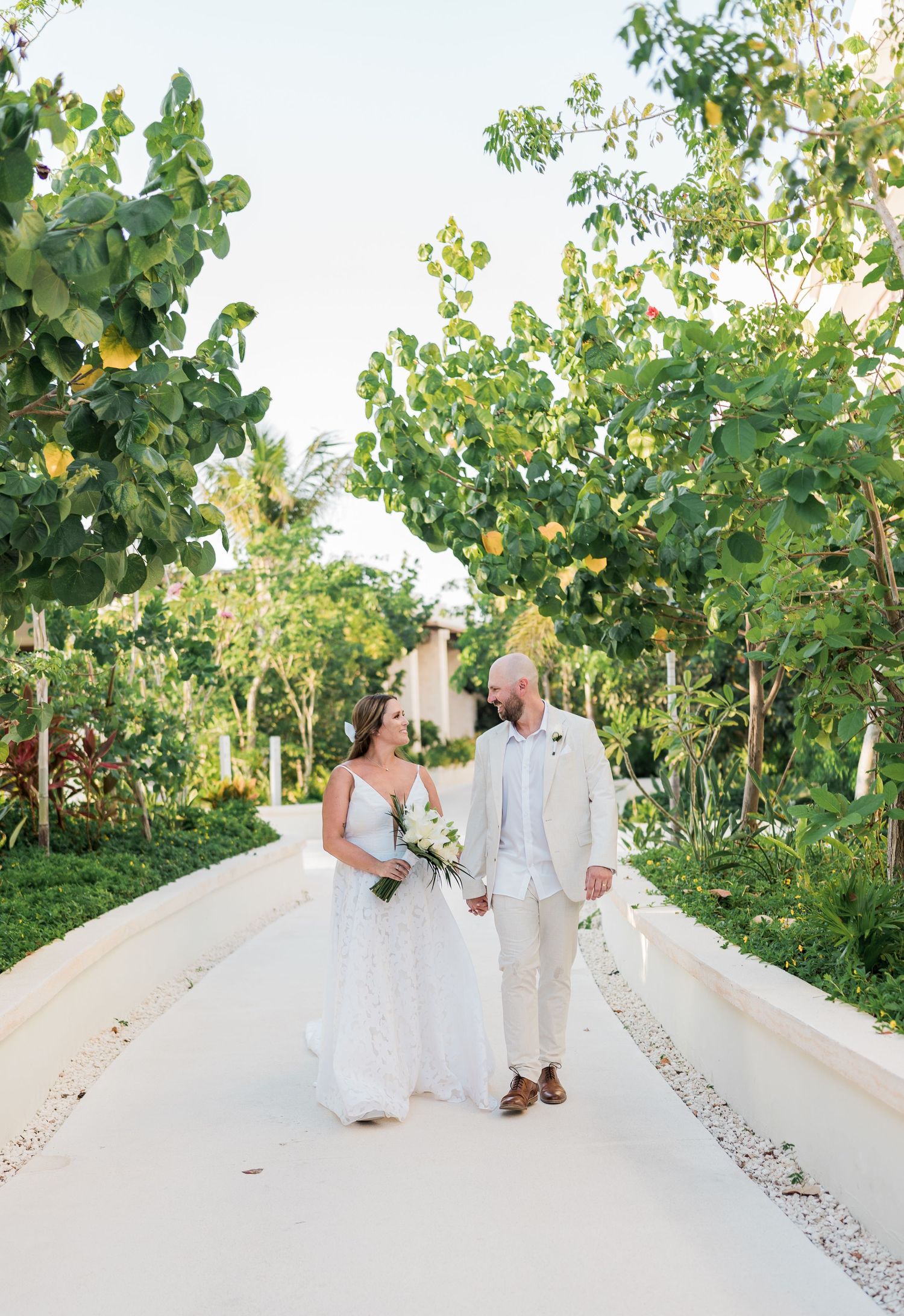 Bride and groom walk hand-in-hand down a lush tropical pathway, bride holding white bouquet in elegant wedding attire.