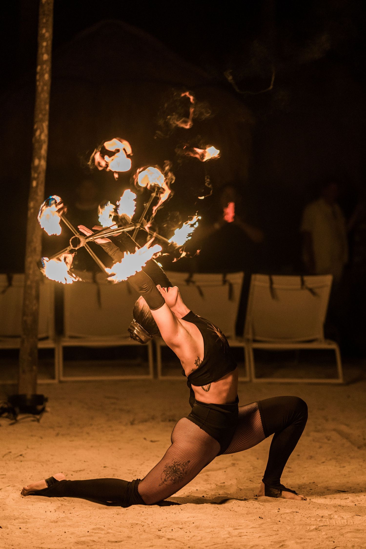 Fire dancer performs stunning backbend on sandy beach at night, spinning flaming poi in dramatic long exposure photography.