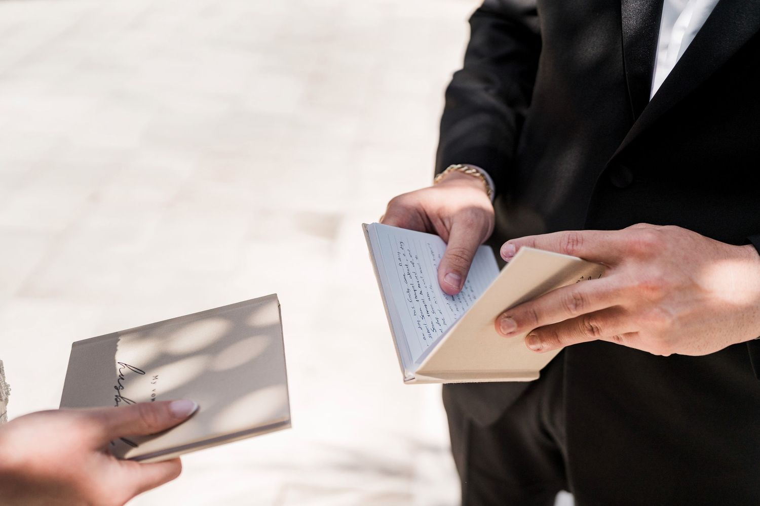 Man in black suit handing over an envelope or card to another person outdoors in bright sunlight.