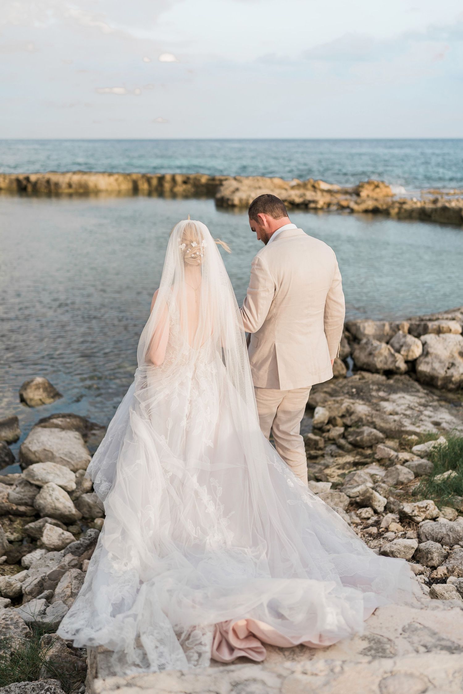 Bride in lace gown and veil stands with groom in tan suit on rocky seaside shore at sunset elopement.