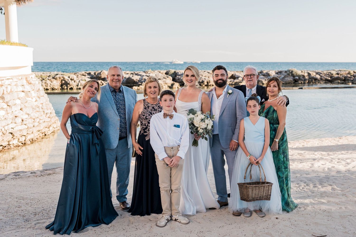 Wedding party poses on a beachside venue with ocean backdrop, featuring bride and groom surrounded by family members in formal attire.