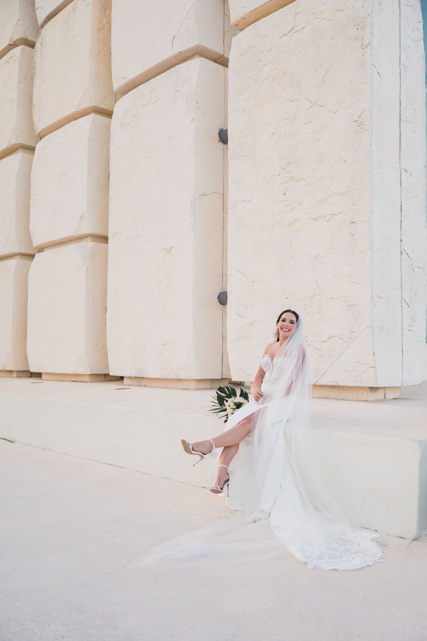 Elegant bride in white lace gown and veil poses with bouquet against grand marble columns.