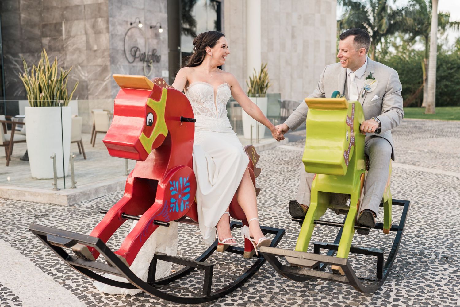 Bride and groom in wedding attire laughing while sitting on colorful rocking horse chairs outdoors.