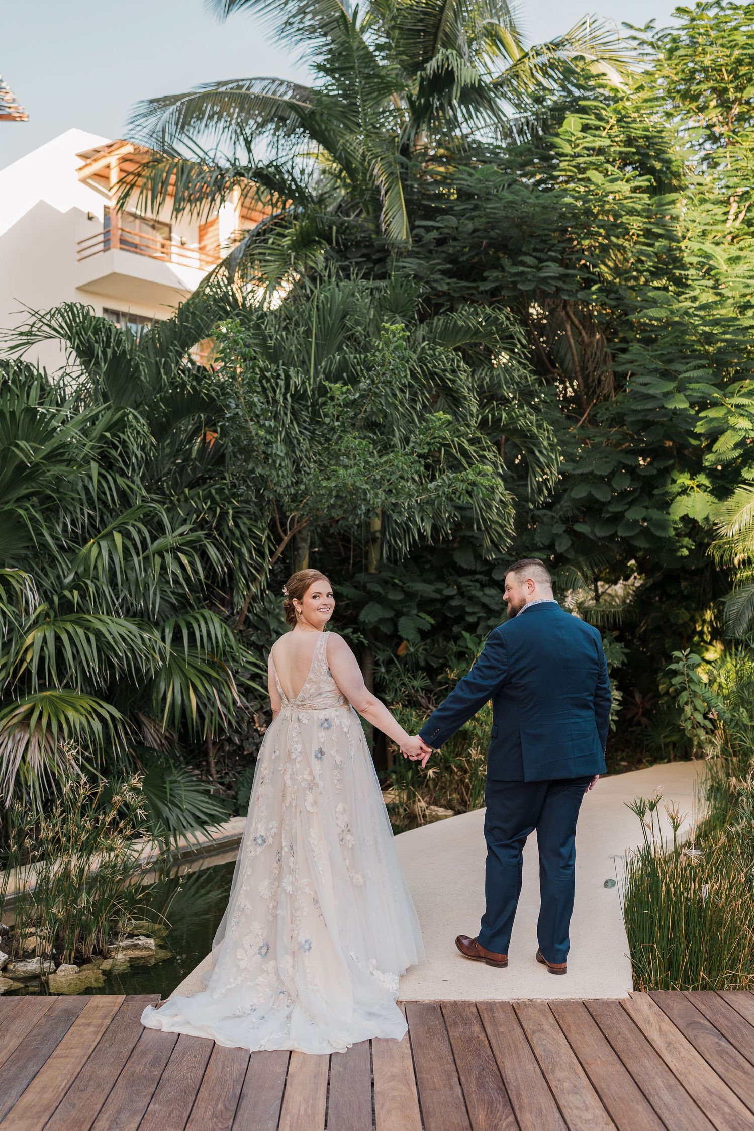 Secrets Akumal Wedding - Bride in floral gown and groom in navy suit hold hands on wooden deck surrounded by lush tropical greenery.