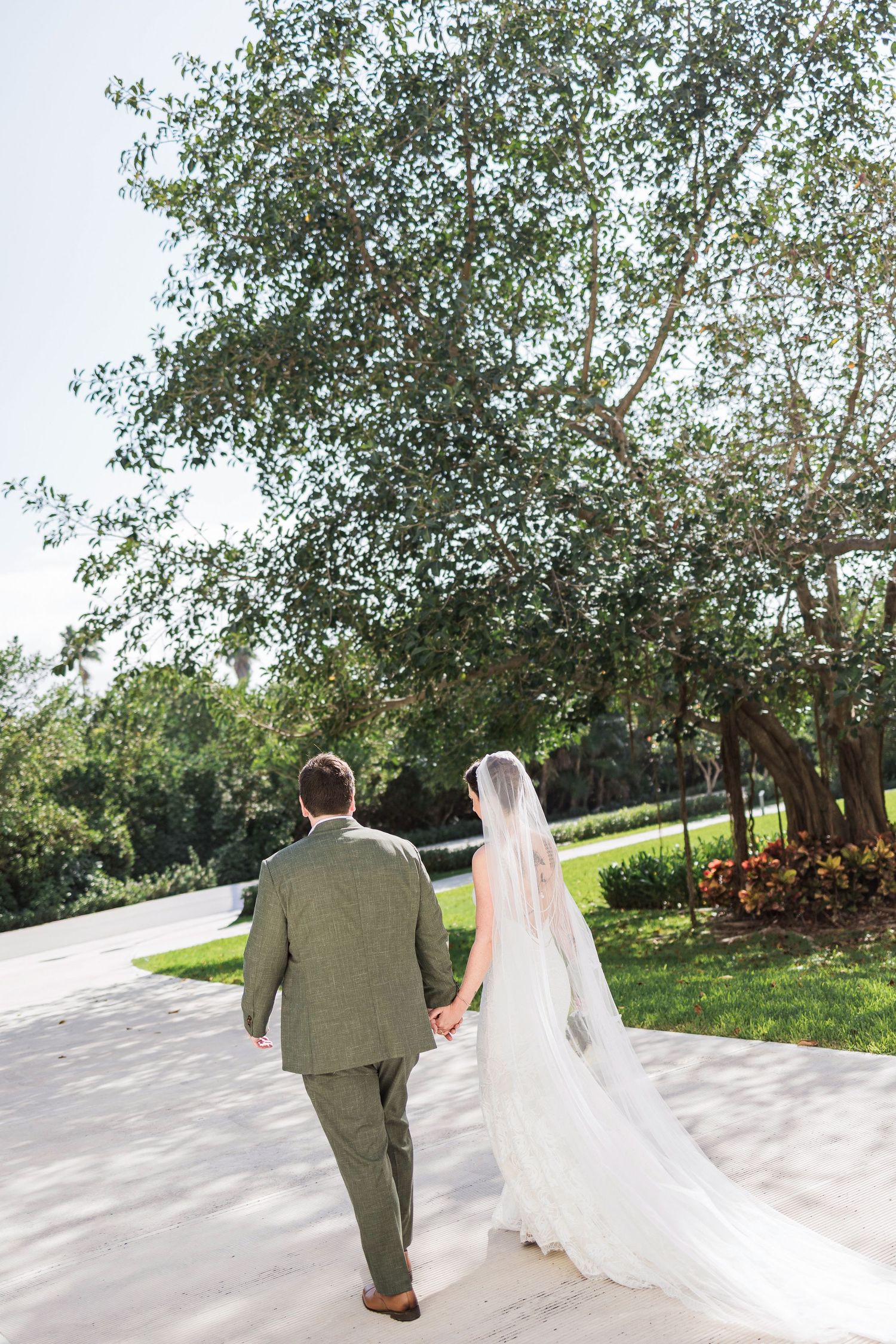 Bride and groom walk hand in hand through lush garden path, bride wearing flowing white gown with long veil.