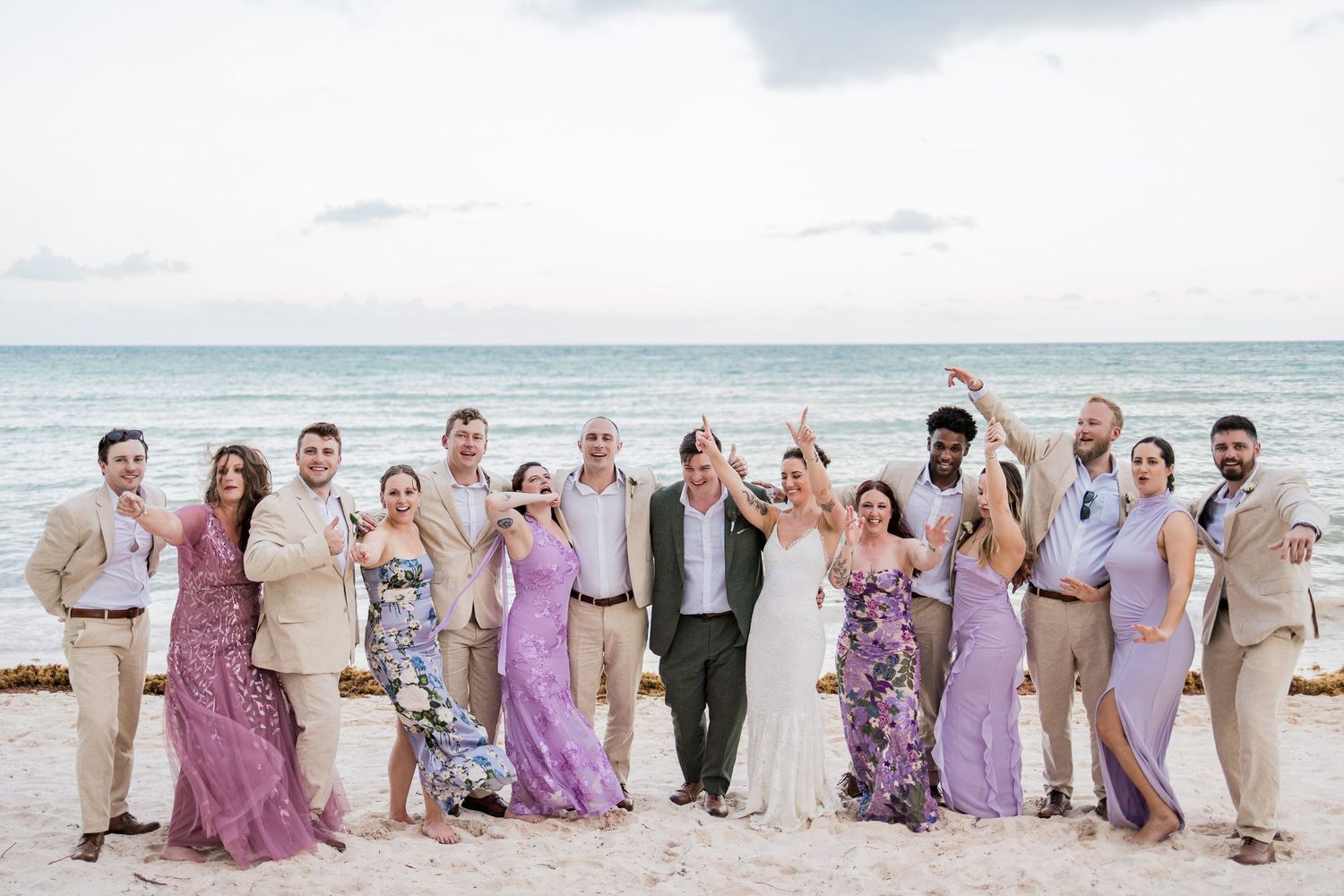 Wedding party celebrating on a sandy beach with ocean backdrop, bridesmaids in purple dresses and groomsmen in tan suits.
