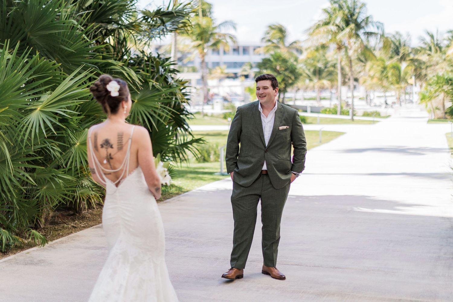 Groom in gray suit smiles at bride in lace backless wedding gown during first look at tropical resort.