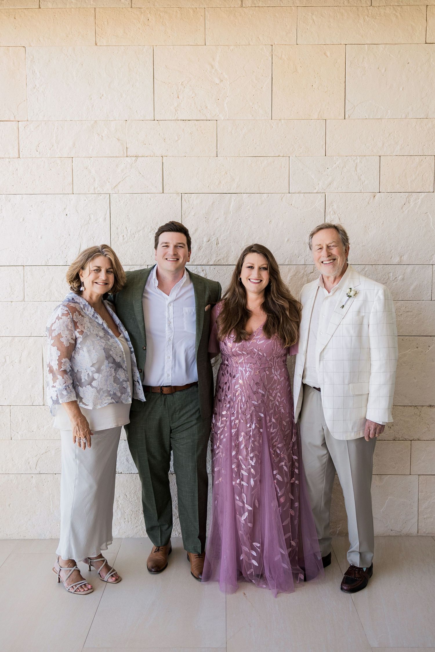 Two couples pose together smiling at a wedding, woman in pink floral gown with family in formal attire.