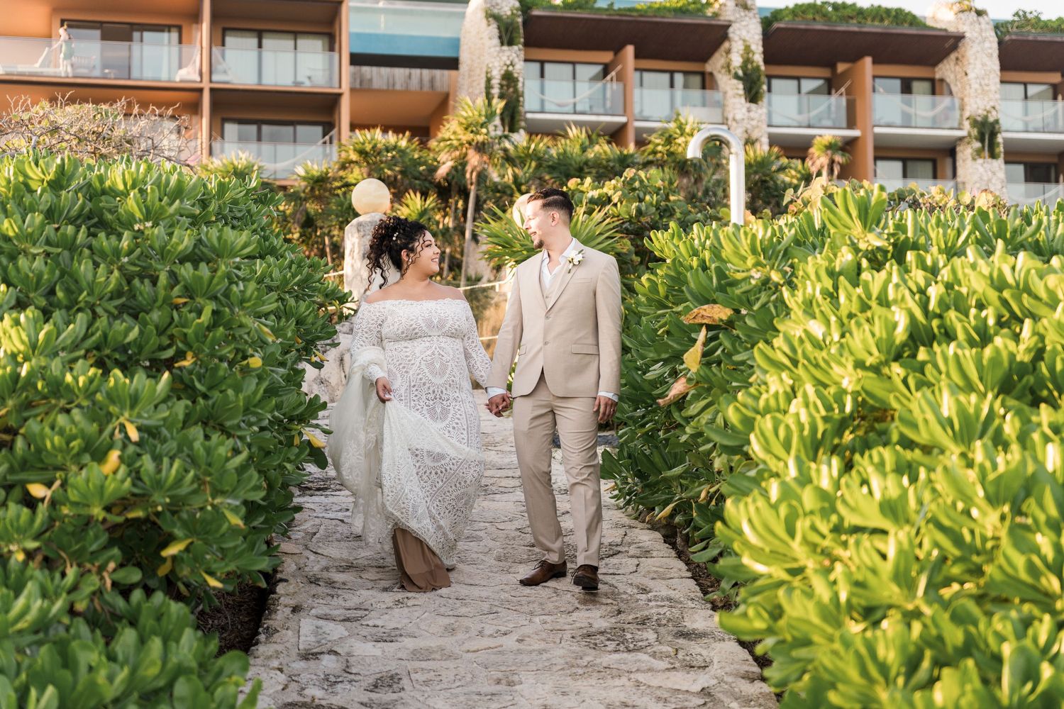 Xcaret Arte Wedding - Bride and groom hold hands on a stone path surrounded by lush greenery with a resort building in the background.