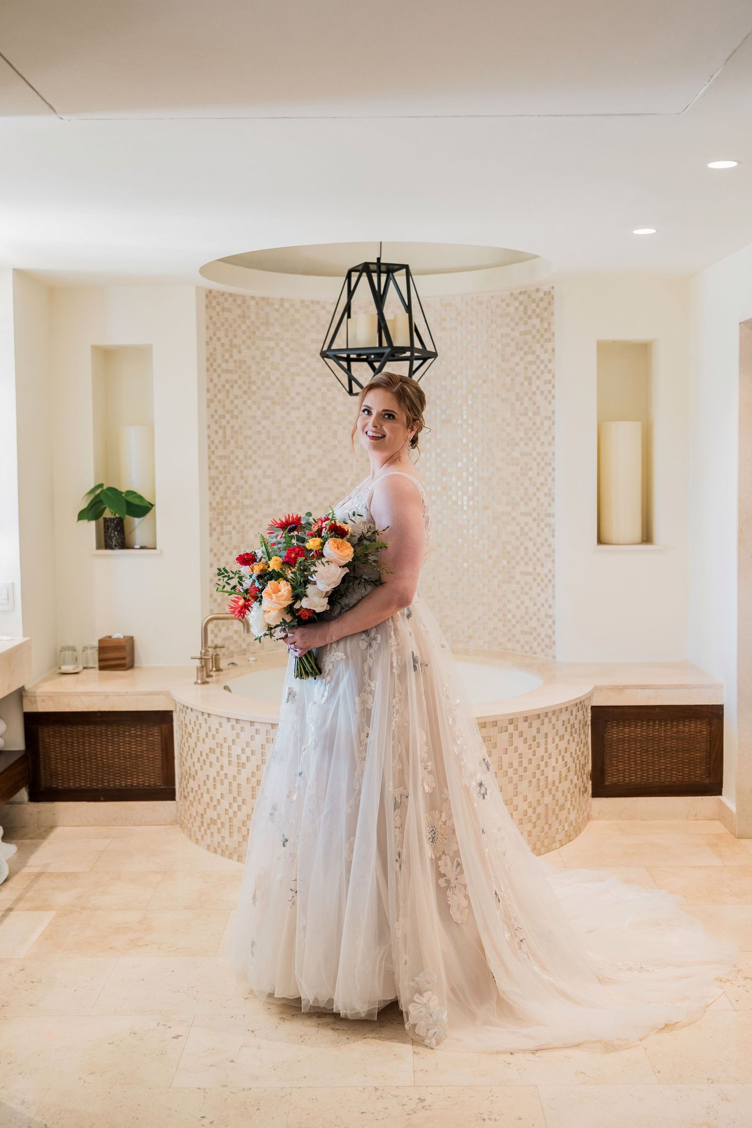 Bride in elegant lace wedding gown holding colorful bouquet stands in luxurious cream-tiled bathroom with soaking tub.