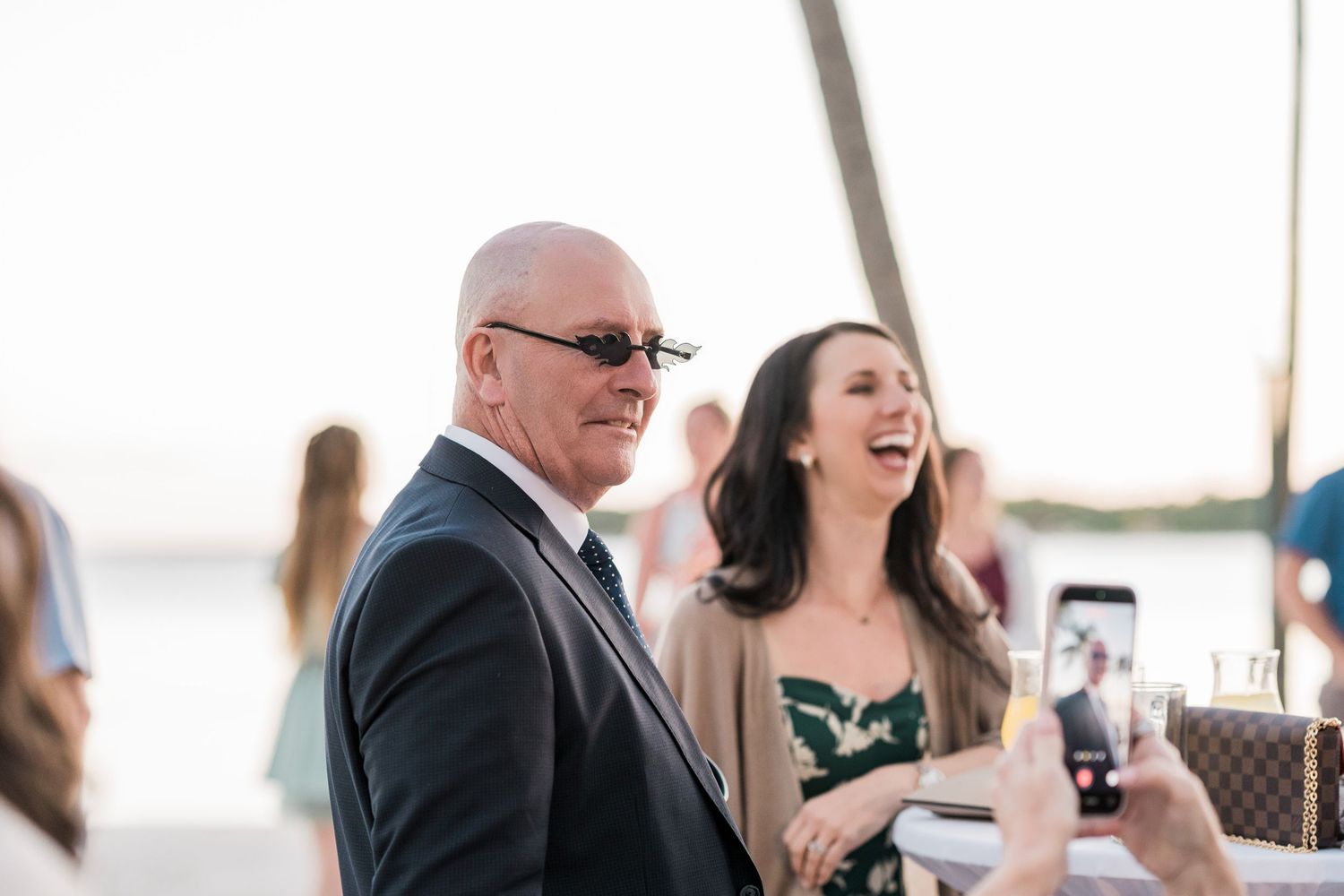 Smiling man in suit and woman in floral dress laughing at an outdoor waterfront event.