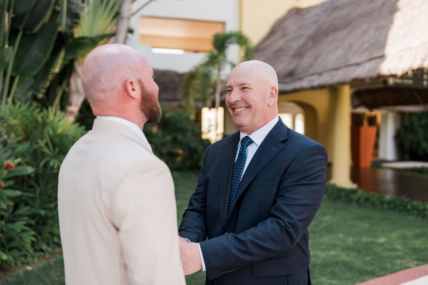 Two bald men in formal suits shaking hands and smiling at an outdoor wedding venue with tropical landscaping.