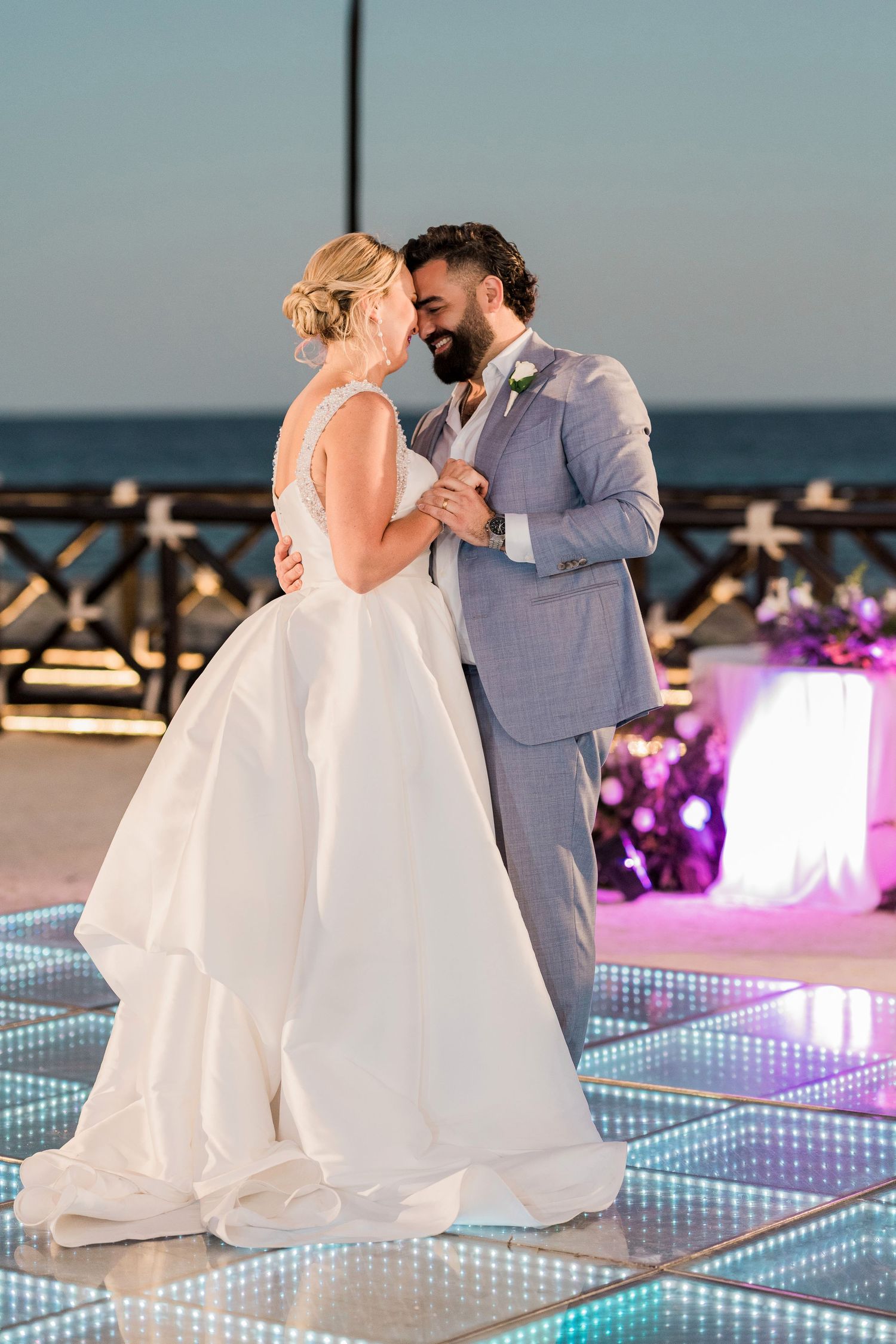 Bride and groom share first dance on illuminated floor at outdoor oceanfront wedding reception at dusk.