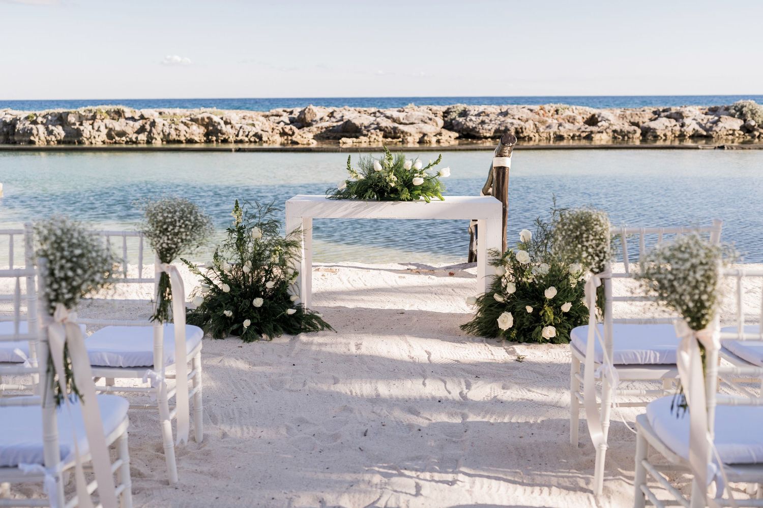 Elegant beachside wedding ceremony setup with white chairs, floral arrangements, and an arch overlooking turquoise ocean waters.