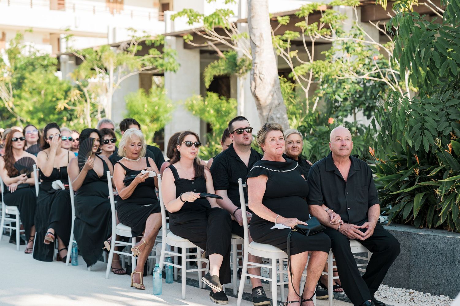Wedding guests dressed in black attire seated in white chairs at an outdoor ceremony surrounded by lush greenery.