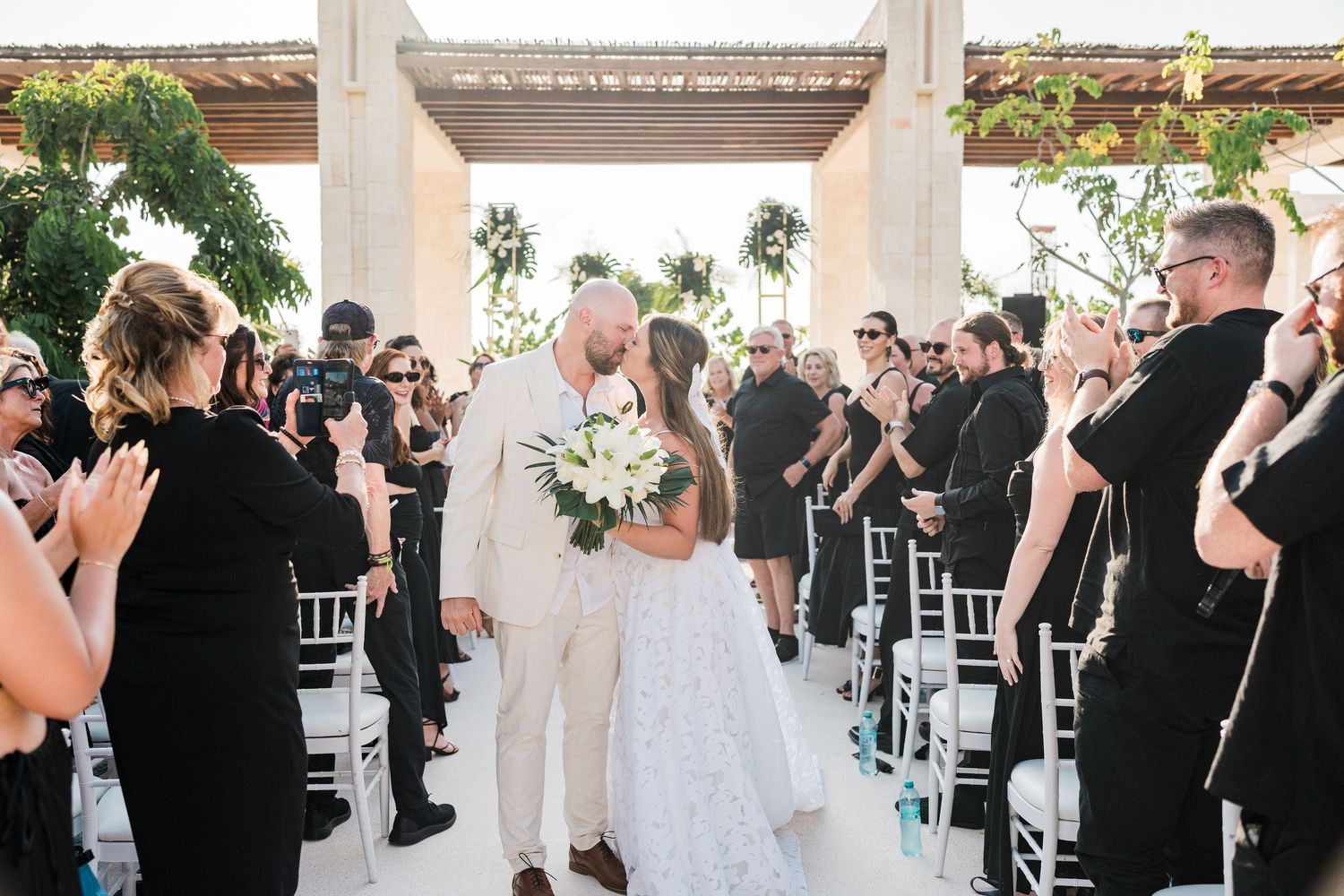 Newlywed couple sharing a kiss at outdoor wedding ceremony while guests in black attire applaud along white chair aisle.