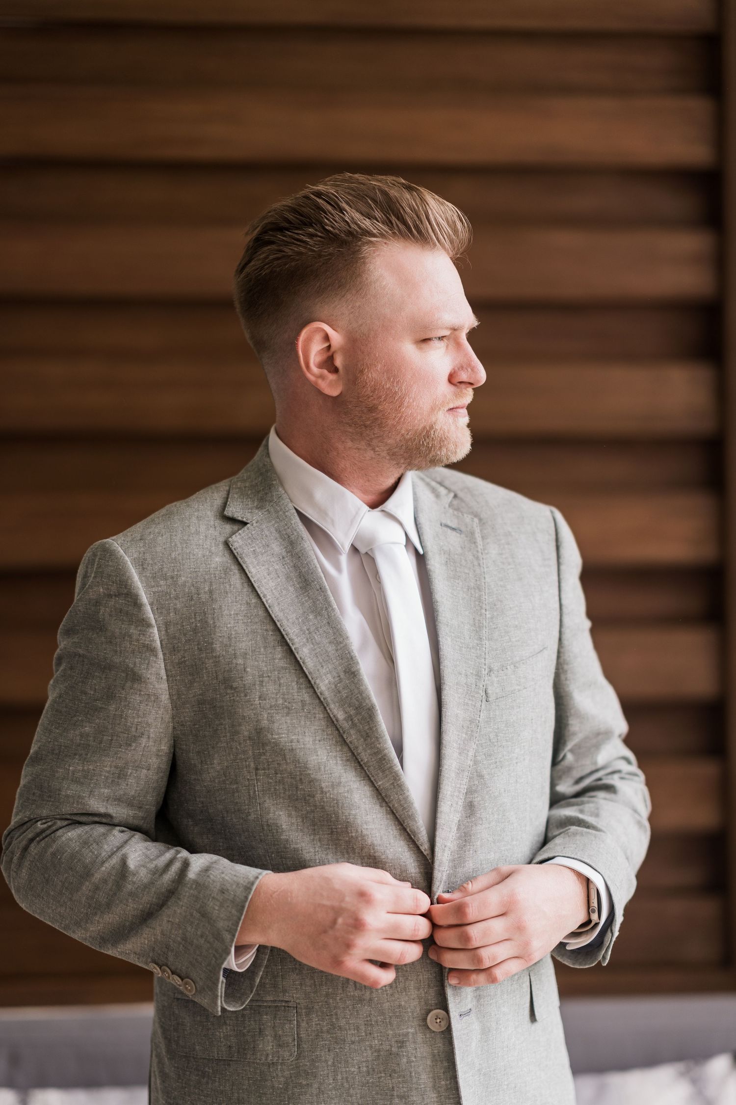 Man in gray suit buttoning jacket, looking sideways, with short styled hair and beard, standing near wooden blinds.